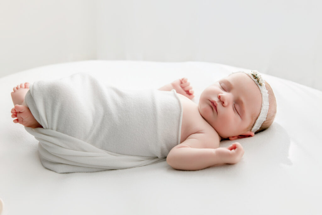 Newborn girl laying on white blanket with flower bow. 